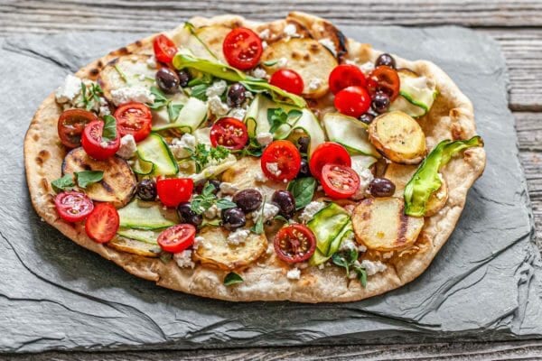 Grilled vegetable pizza topped with halved tomatoes, black olives, cheese, zucchini ribbons and sliced potatoes. The pizza is on a slate board.