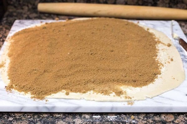 Dough for the best cinnamon rolls rolled out and topped with cinnamon and sugar mixture. A rolling pin is behind the dough.