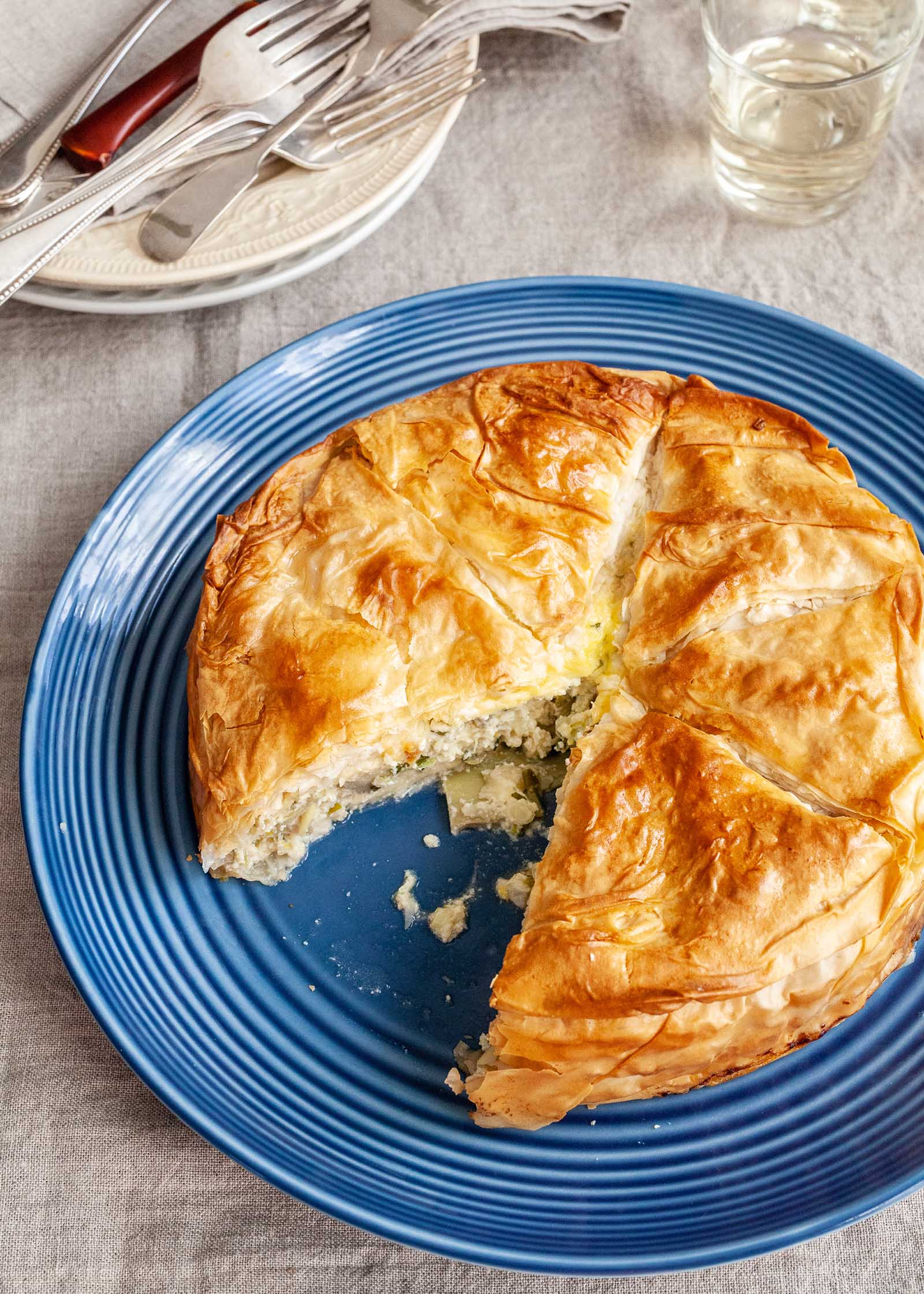 A flaky spring pie is on a blue plate and one slice is missing. The cheesy artichoke filling is visible on the plate. A grey tablecloth is under the plate and a stack of white plates and pile of forks is in the upper left corner. A glass of white wine is in the upper right corner.