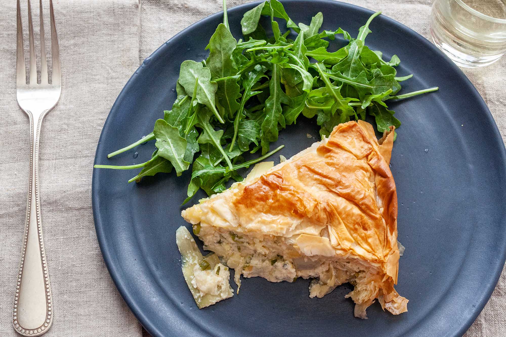 Overhead horizontal view of a blue plate with a slice of cheese pie with artichokes on it. The filling is slightly spilling out of the slice. A green arugula salad is behind the pie on the plate. A silver fork is to the left of the plate and a glass is in partial view in the upper right corner.