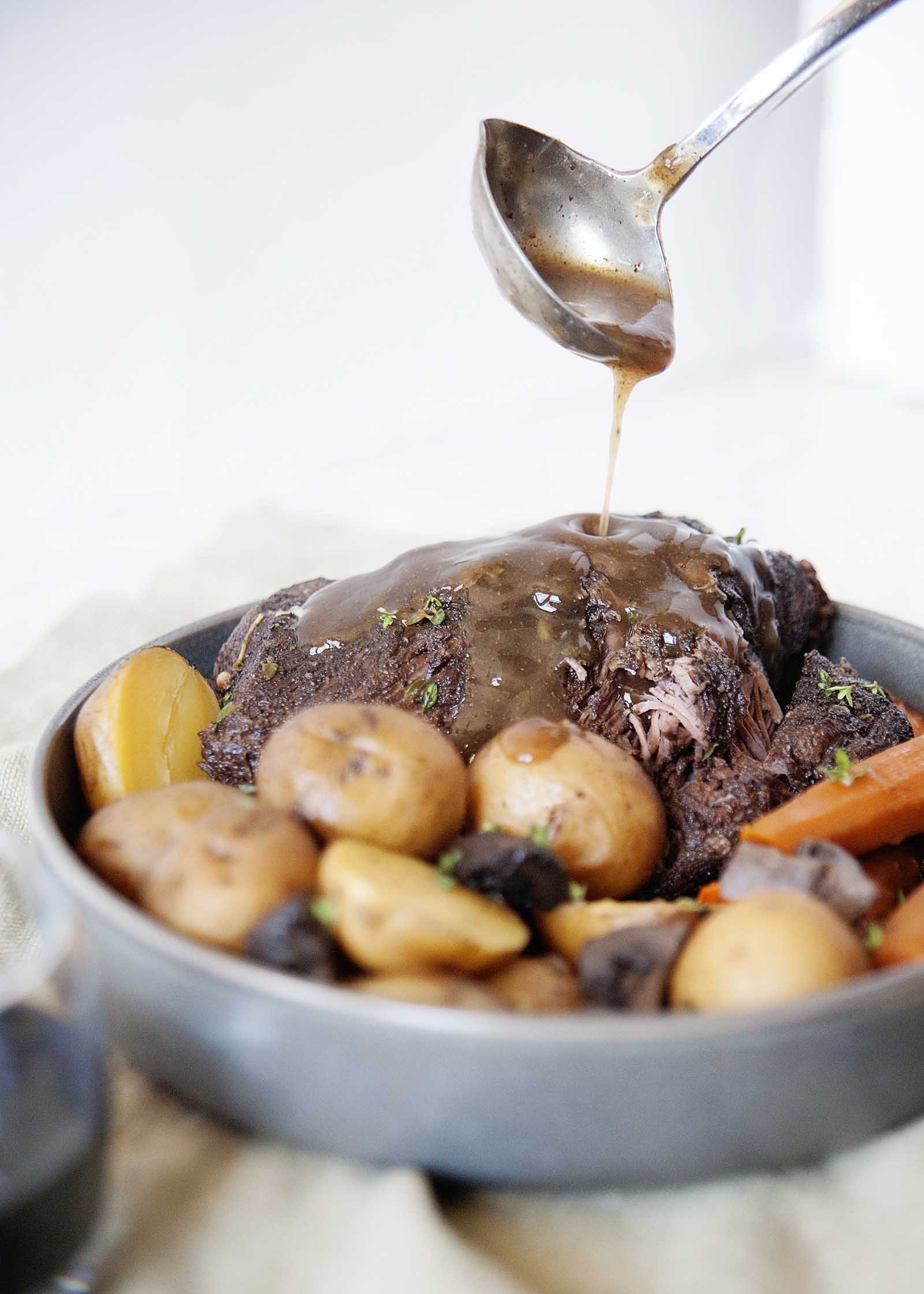 Ladling gravy over a platter full of pot roast, potatoes and carrots. 