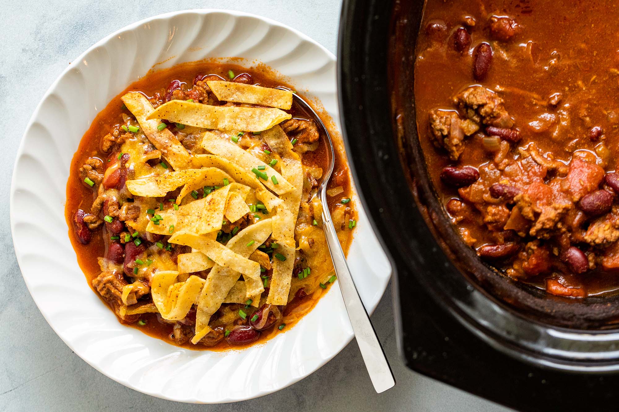 Easy Slow Cooker Beef and Bean Chili topped with fried tortilla stips in a white bowl. A crock pot is to the right of the bowl.