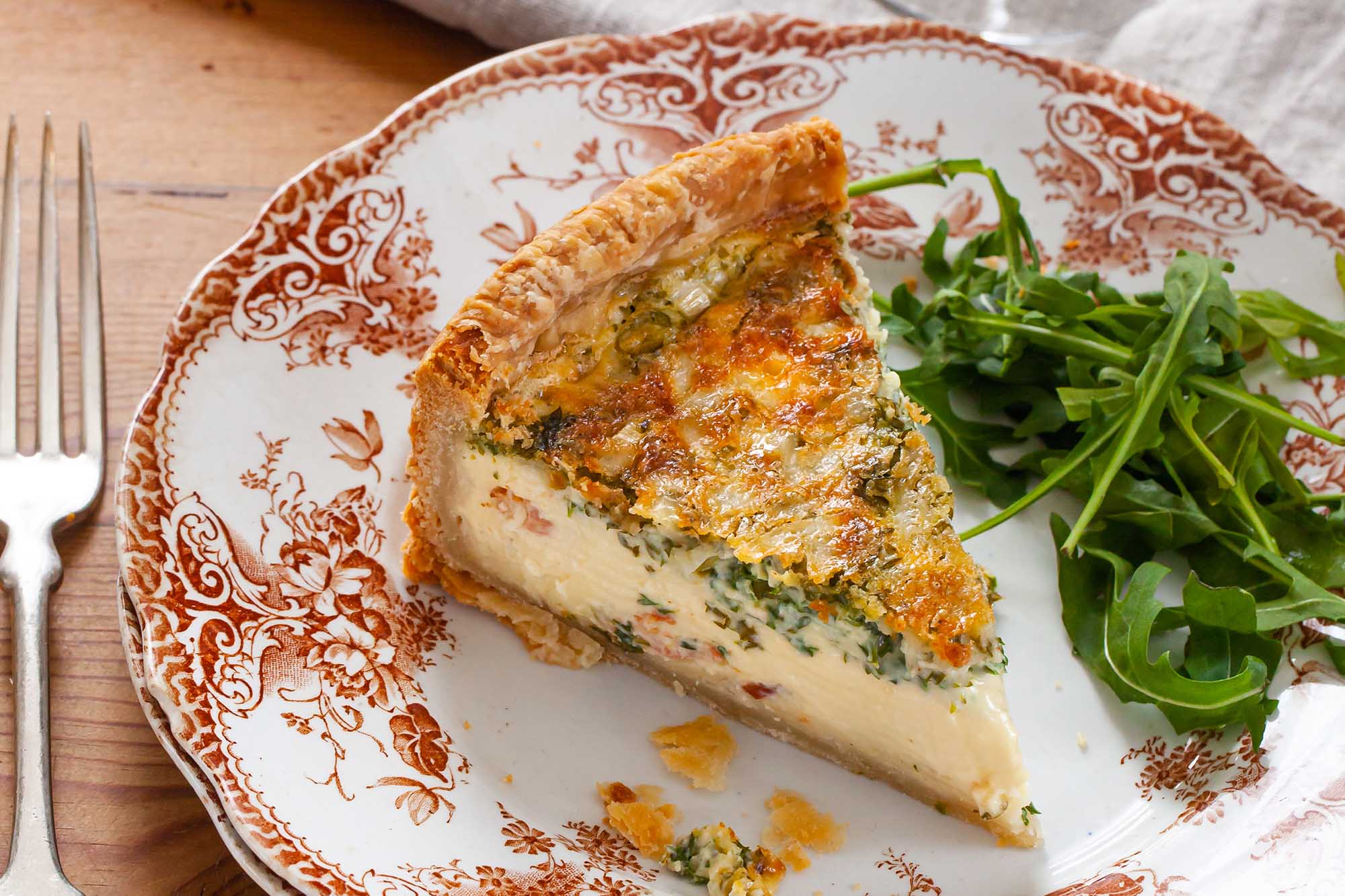 A slice of best deep dish quiche on a decorative china plate. A handful of arugula is to the right of the quiche. A fork is to the left of the plate and a table linen is above the plate.