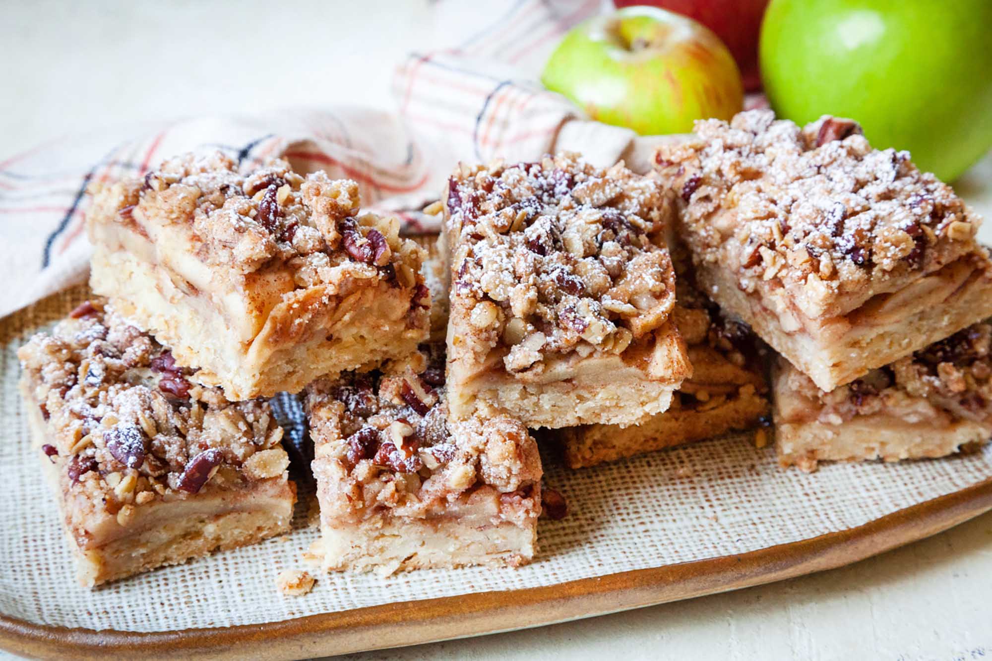 Apple pie bars with pecan oat crumb topping stacked on a platter with apples behind them.