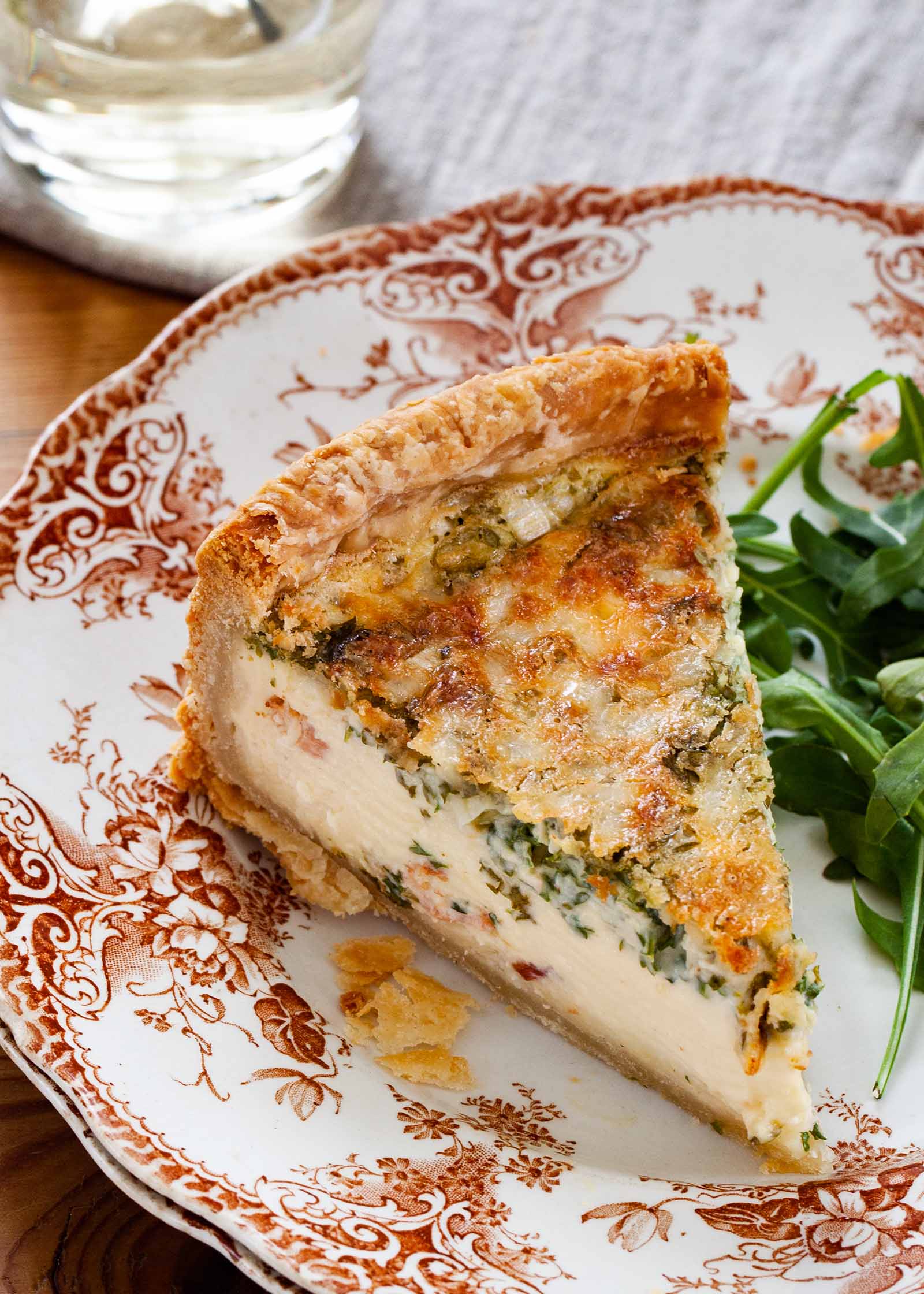 A slice of best deep dish quiche on a decorative china plate. A handful of arugula is to the right of the quiche and a table linen and glass are above the plate.