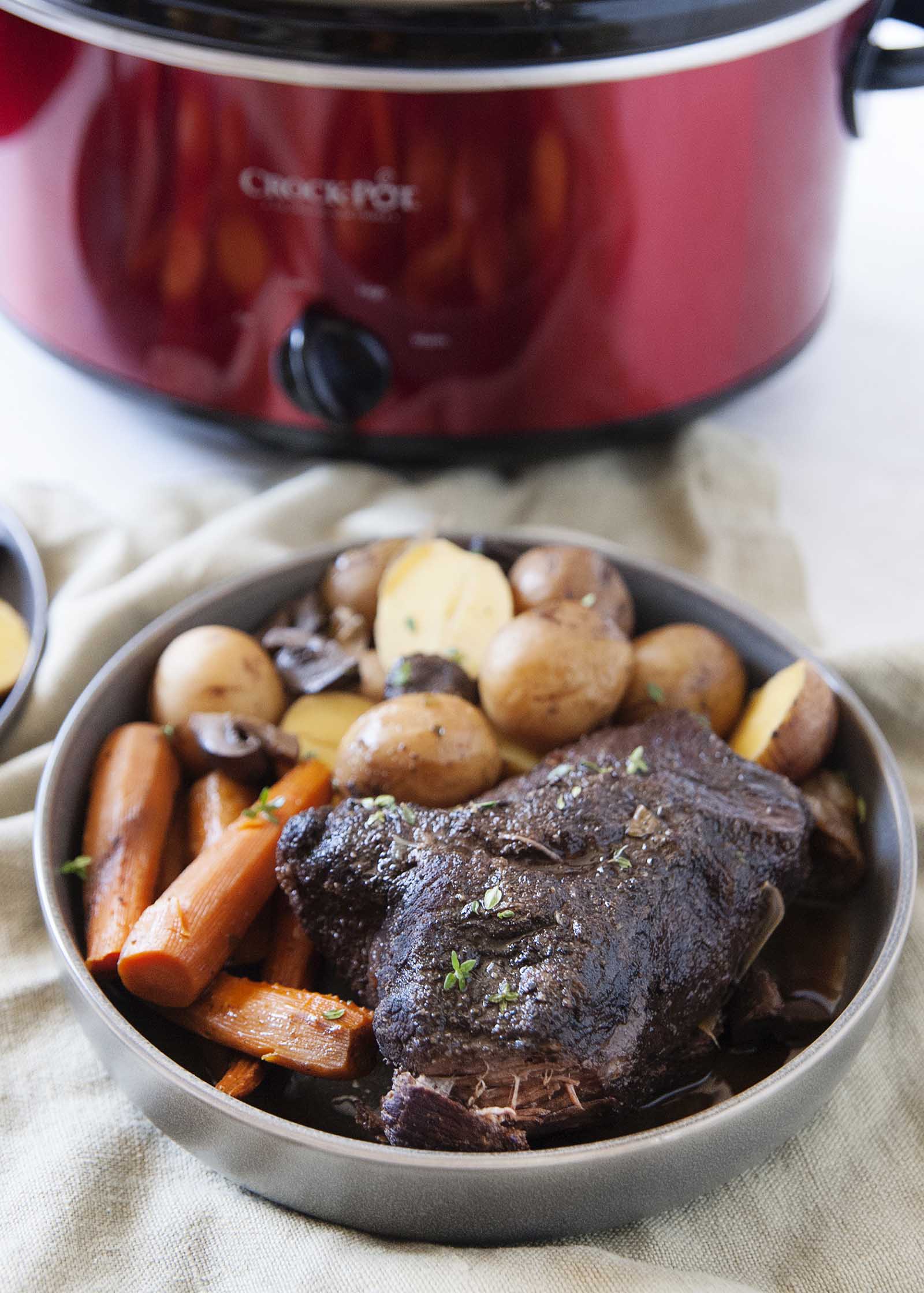 A grey platter with slow cooker pot roast and gravy and carrots and potatoes in front of a red slow cooker.